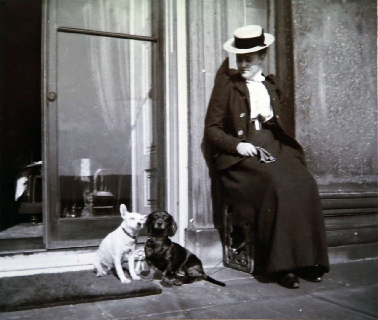 Black and white home photograph of a Victorian lady wearing an overcoat, long skirt, shirt and straw hat, sat out on the Terrace looking down at her two Sausage dogs.