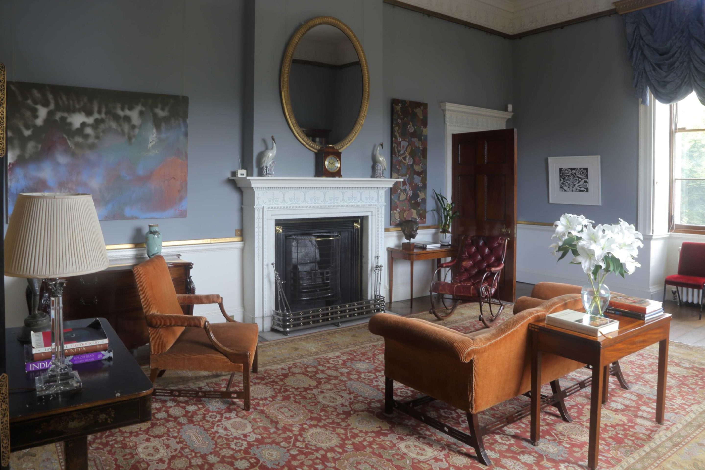 A large room with grey-blue walls hung with several pieces of contemporary art. There is a brown suite of furniture and a leather rocking chair.