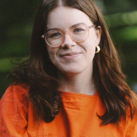 A person with mid length brown hair, smiling warmly against a lush green background. The person wears light orange glasses, gold earrings, nose ring and a bright orange top.