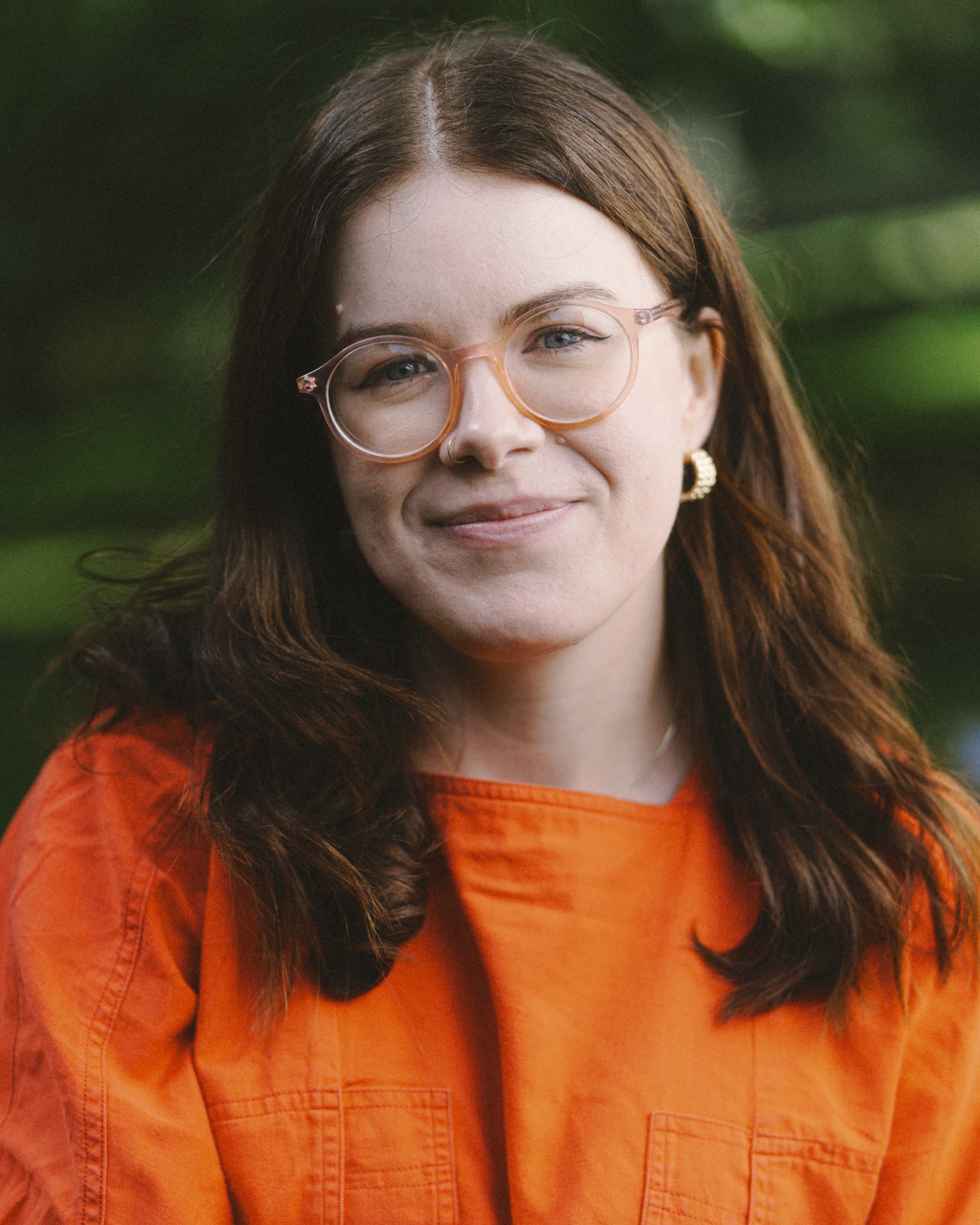 A person with mid length brown hair, smiling warmly against a lush green background. The person wears light orange glasses, gold earrings, nose ring and a bright orange top.