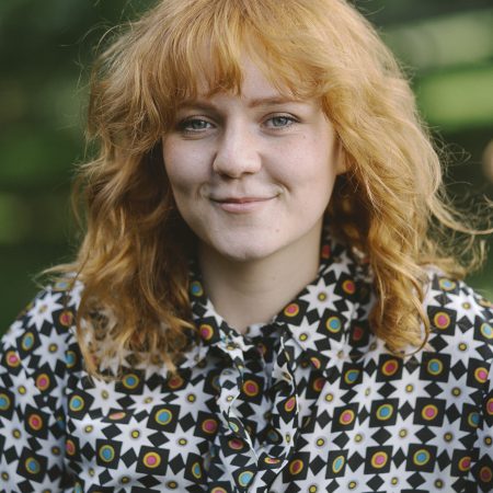 A smiling ginger person in her 20's with curly ginger hair and a brightly patterned shirt