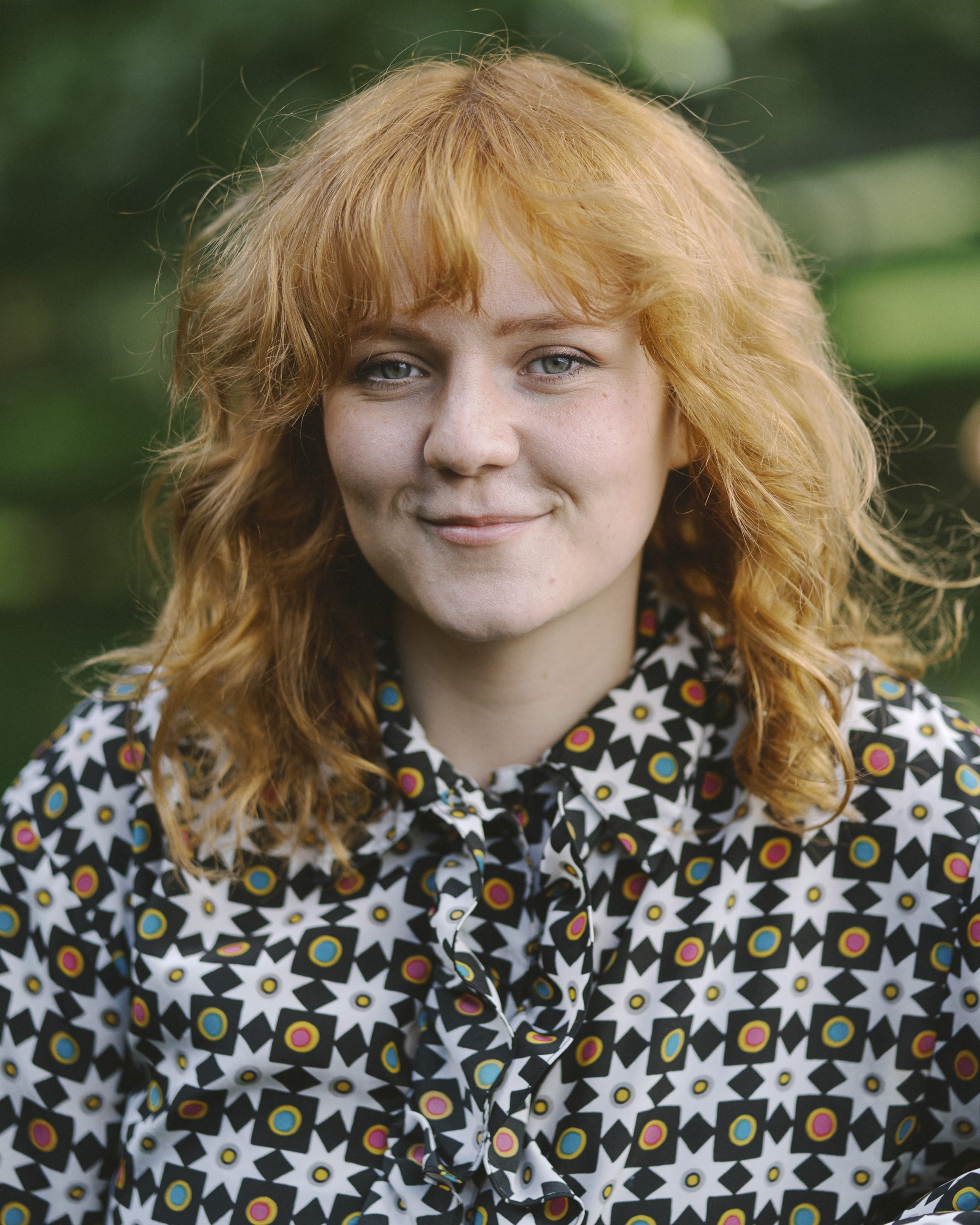 A smiling ginger person in her 20's with curly ginger hair and a brightly patterned shirt