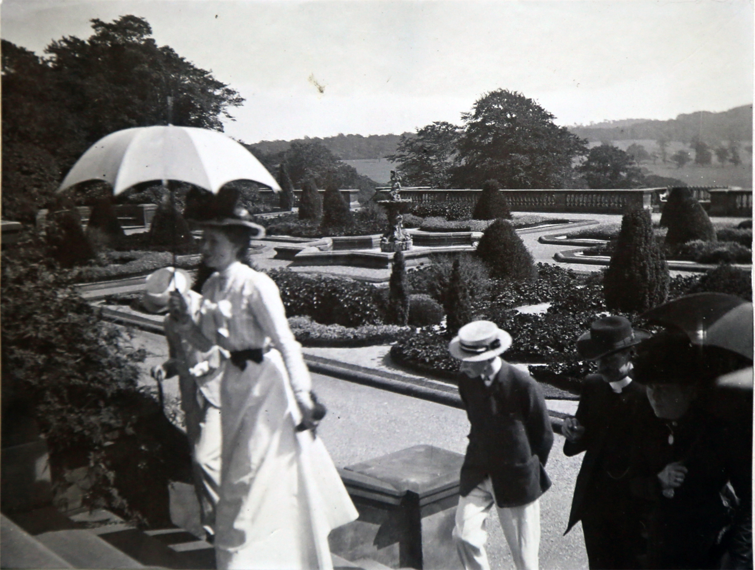 Black and white home photograph of well dressed victorians walking up the steps of the Terrace. A women in a long light coloured dress wears a hat and holds an umbrella, as gentleman walk up the stairs behind her.