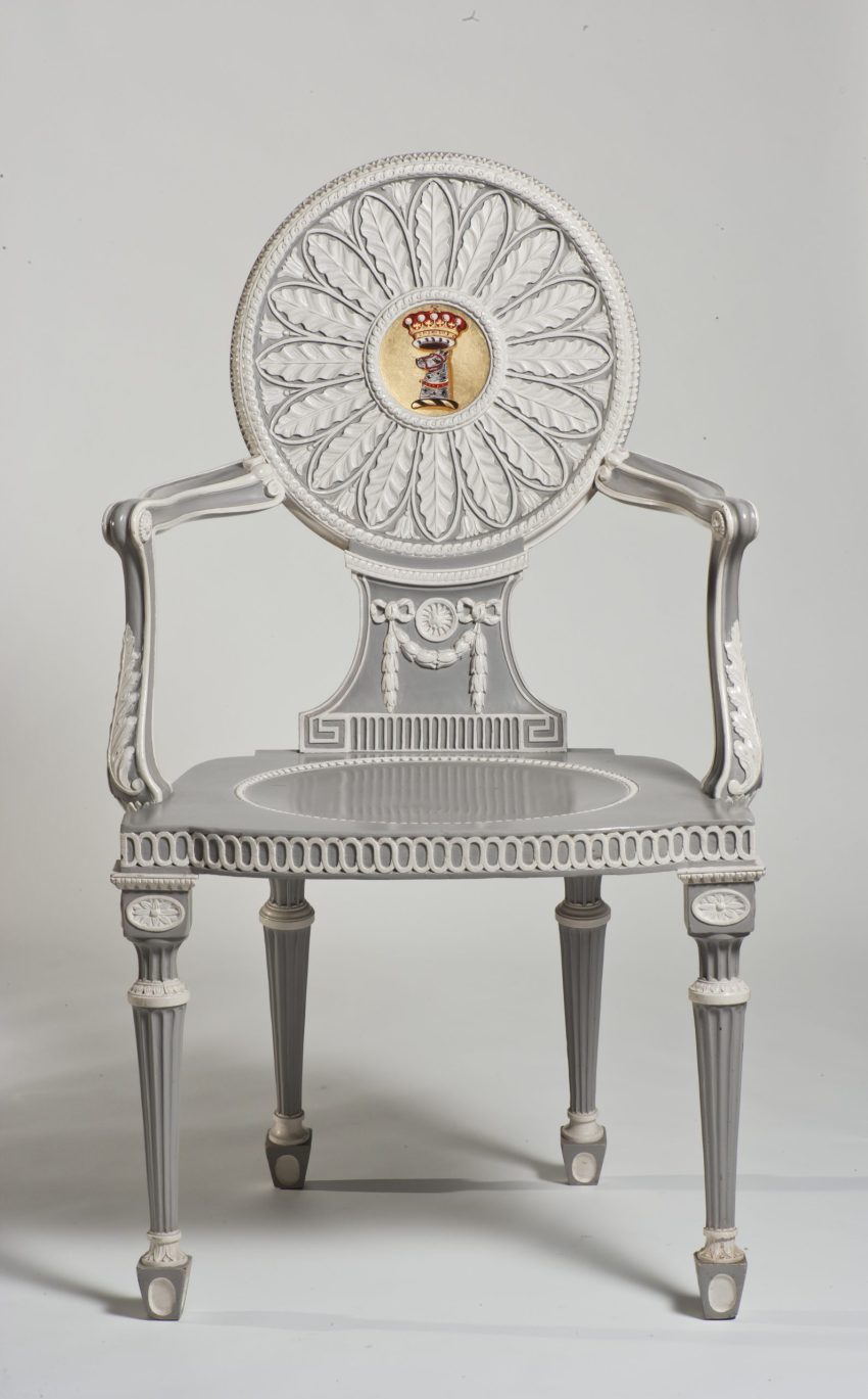 An intricately carved grey and white painted chair, with a gold crest in the centre of the seat back.