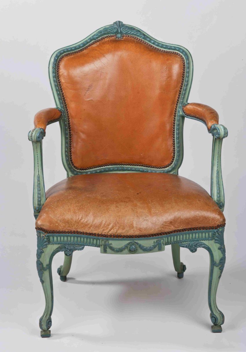 An arm chair with a decorative carved and blue-painted frame, and tan leather seat an arm cushions.