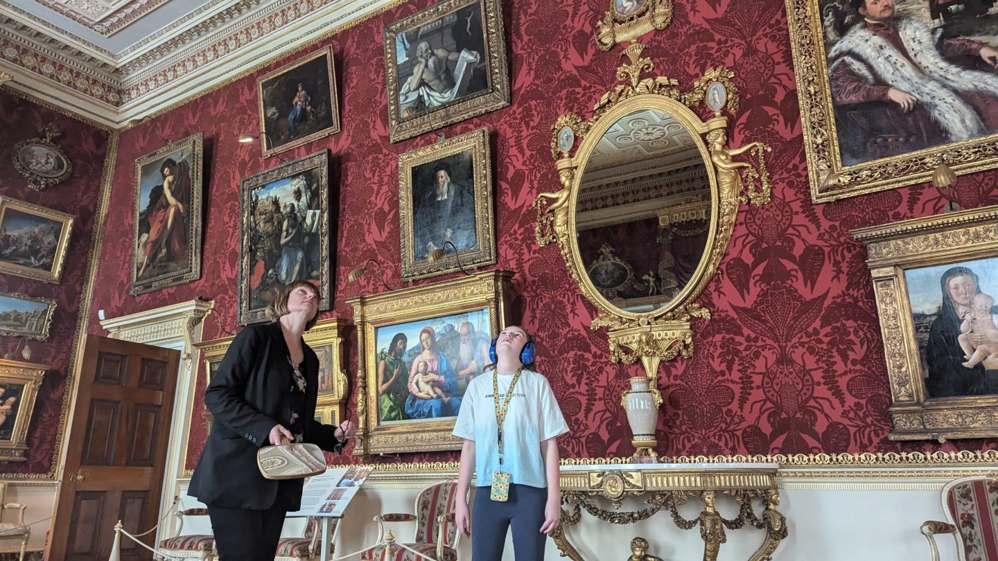 Two people stand in the middle of the grand gallery looking up at the intricate Robert Adam ceiling. The walls are covered in deep red damask, adorned with an array of gilded-framed paintings. A large ornate mirror reflects the opulent surroundings, and a decorative console table holds a classical urn. One person holds a mirror, while the other wears ear defenders and a sunflower lanyard, fully immersed in the experience.