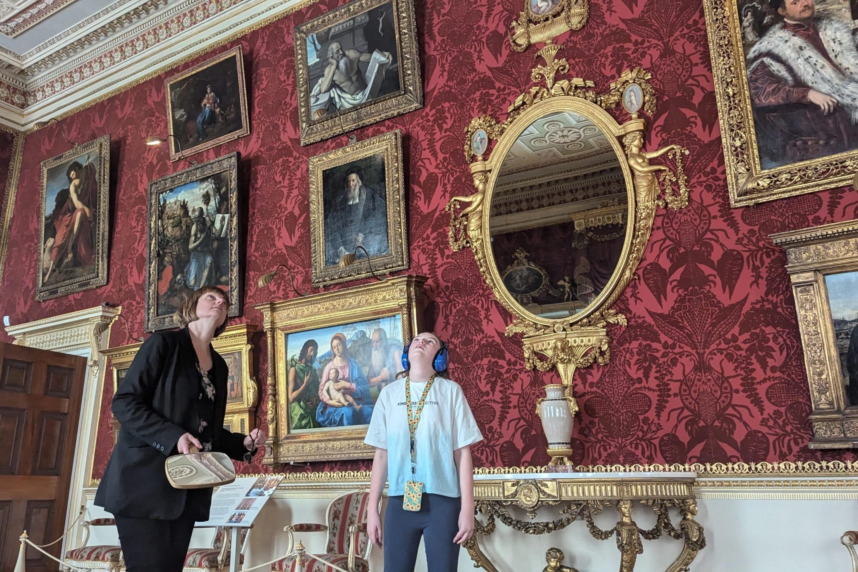 Two people stand in the middle of the grand gallery looking up at the intricate Robert Adam ceiling. The walls are covered in deep red damask, adorned with an array of gilded-framed paintings. A large ornate mirror reflects the opulent surroundings, and a decorative console table holds a classical urn. One person holds a mirror, while the other wears ear defenders and a sunflower lanyard, fully immersed in the experience.