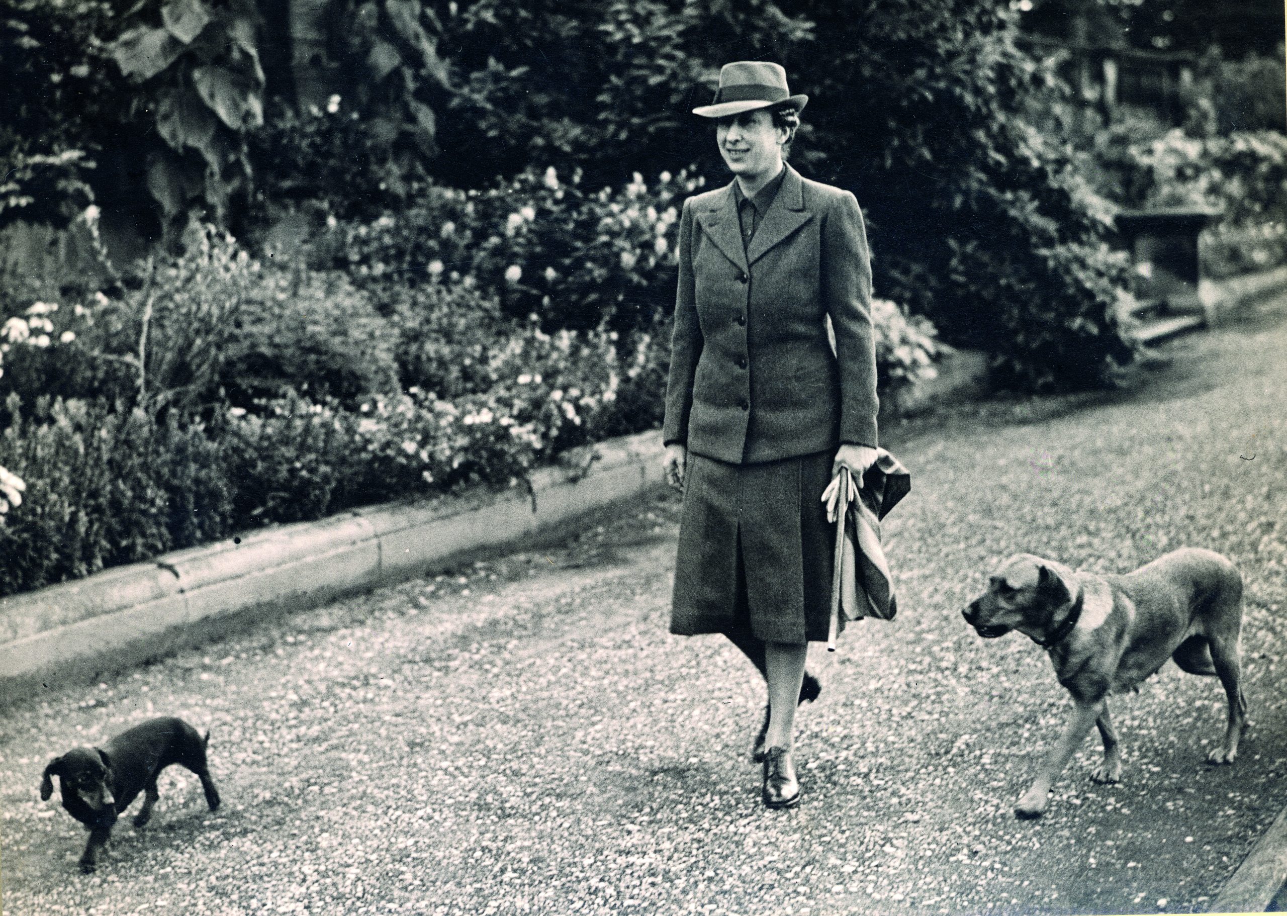 Black and white photograph of Princess Mary walking her two dogs, a black sausage dog and a red labrador, on the Terrace. Princess Mary wears a matching tweed shirt, blazer and fedora hat.