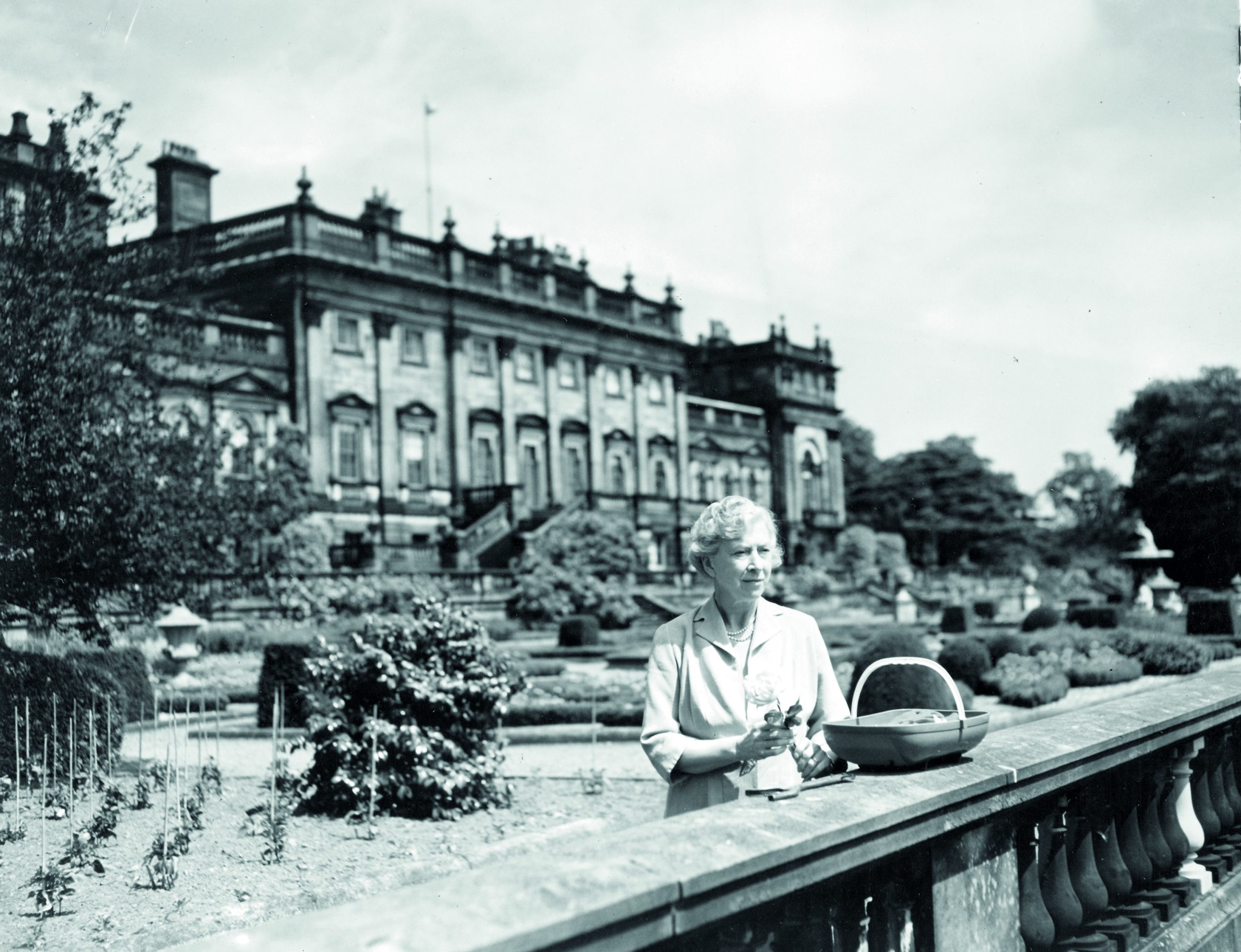 Black and white photography of Princess Mary holding cuttings she's taken from her Terrace Garden. The House stands proudly in the background softly out of focus, with beans and peas growing on the Terrace behind her.