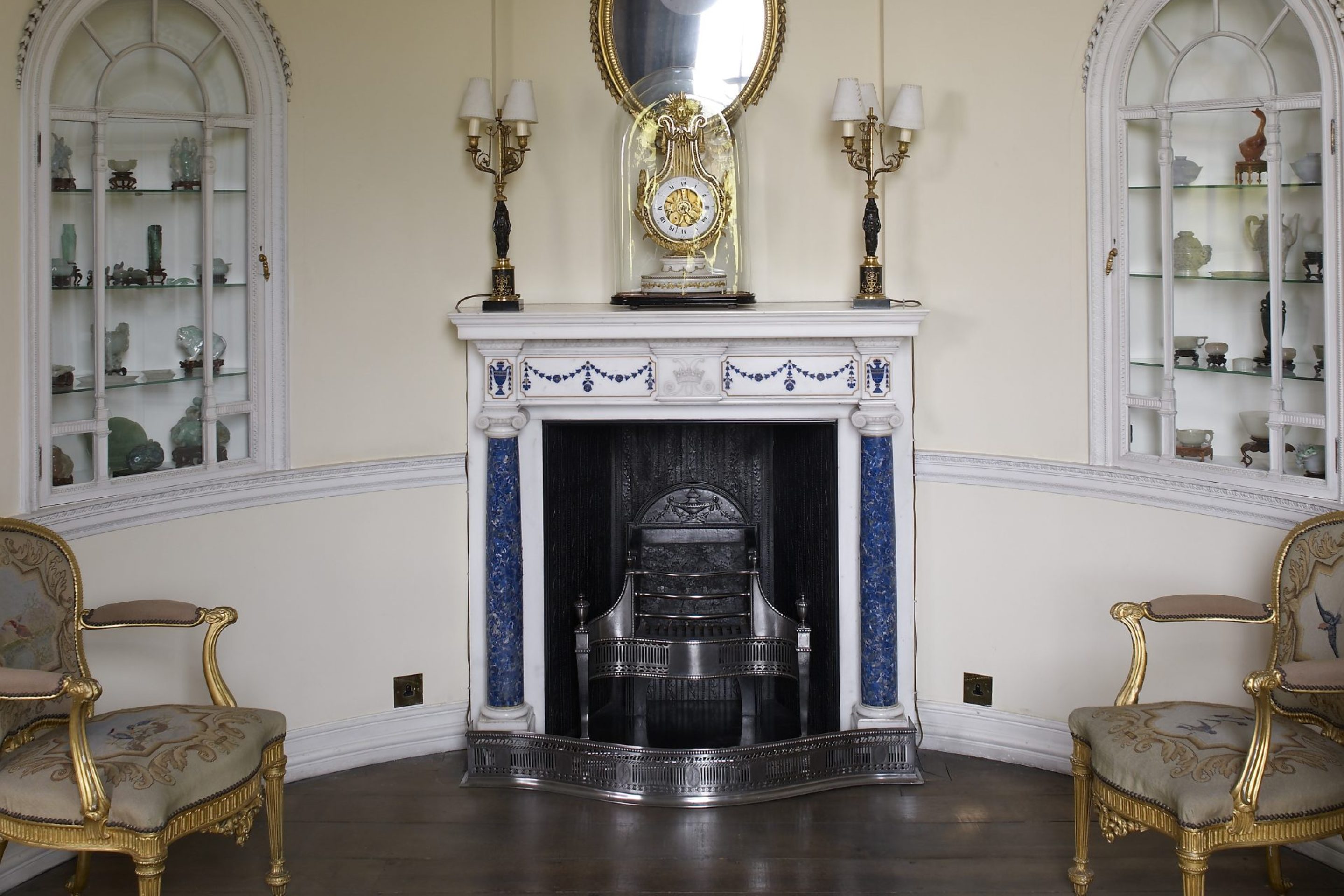 An alcove with a fireplace in the centre and two arched display cabinets to the left and right. The mantlepiece is decorated with lapiz lazuli columns and detailing, and a large lyre-shaped clock sits on top.