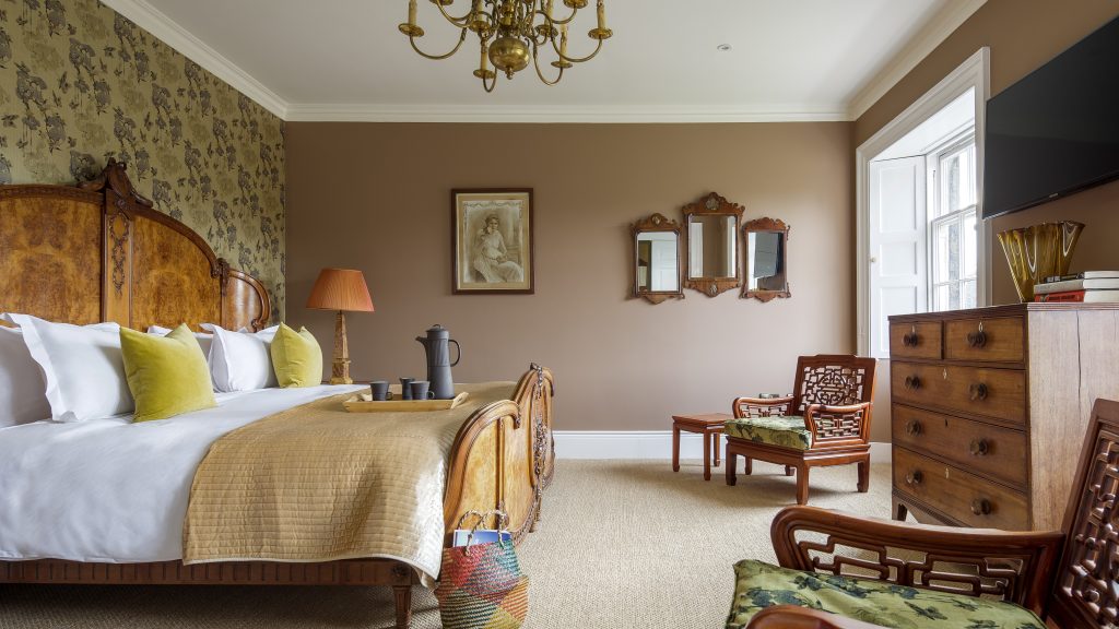 A luxurious bedroom featuring a grand wooden bed with intricate carvings, dressed in crisp white linens, mustard yellow pillows, and a golden quilt. The walls are painted a warm brown with floral-patterned wallpaper on an accent wall. The room is decorated with vintage furniture, including a wooden chest of drawers, antique mirrors, and Chinese-style chairs with green floral cushions. A central chandelier hangs from the ceiling, adding elegance, while natural light streams in through a large window.