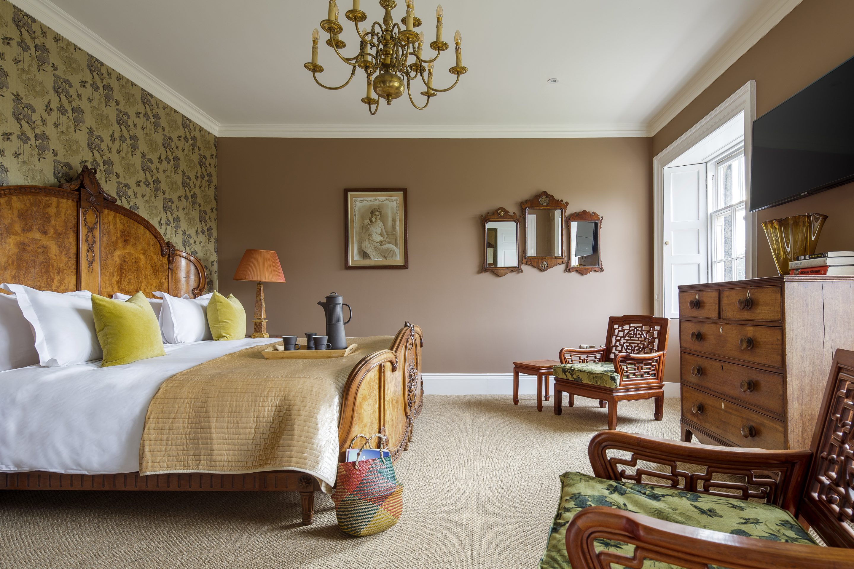 A luxurious bedroom featuring a grand wooden bed with intricate carvings, dressed in crisp white linens, mustard yellow pillows, and a golden quilt. The walls are painted a warm brown with floral-patterned wallpaper on an accent wall. The room is decorated with vintage furniture, including a wooden chest of drawers, antique mirrors, and Chinese-style chairs with green floral cushions. A central chandelier hangs from the ceiling, adding elegance, while natural light streams in through a large window.