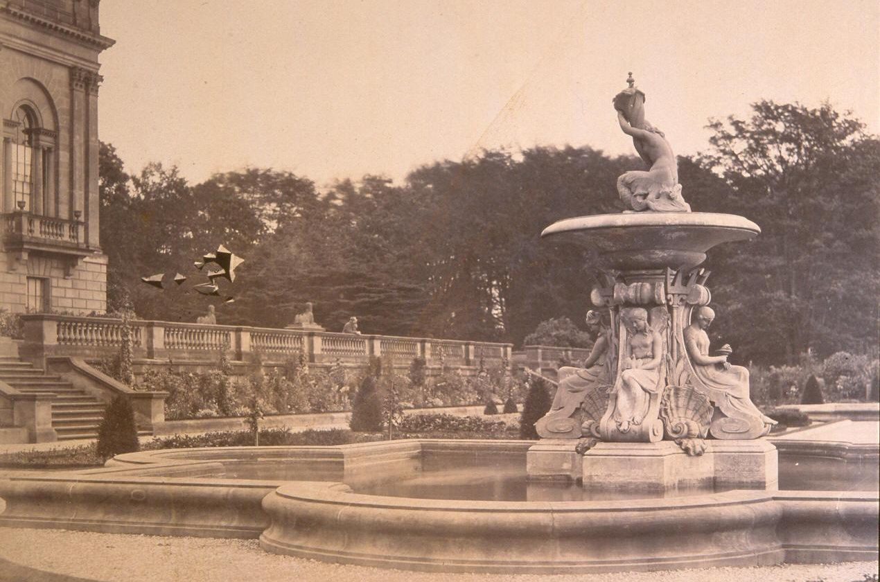 Sepia photograph of the original water fountain that stood in the middle of the Victorian Terrace captured in the mid 1800s. The water fountain features an intricate design of four greek goddesses sitting below a large plate with another goddess on top holding a large flute.