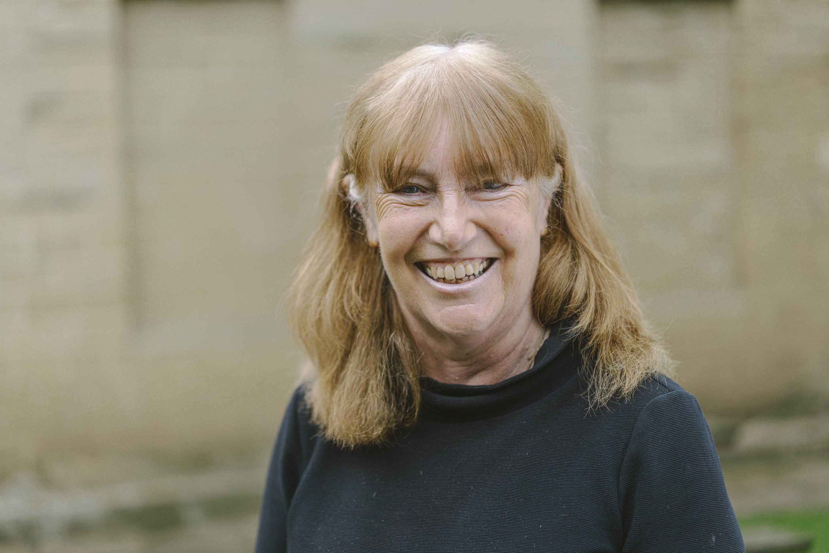 A smiling person with light brown shoulder length and full fringe hair, wearing a black round neck top.