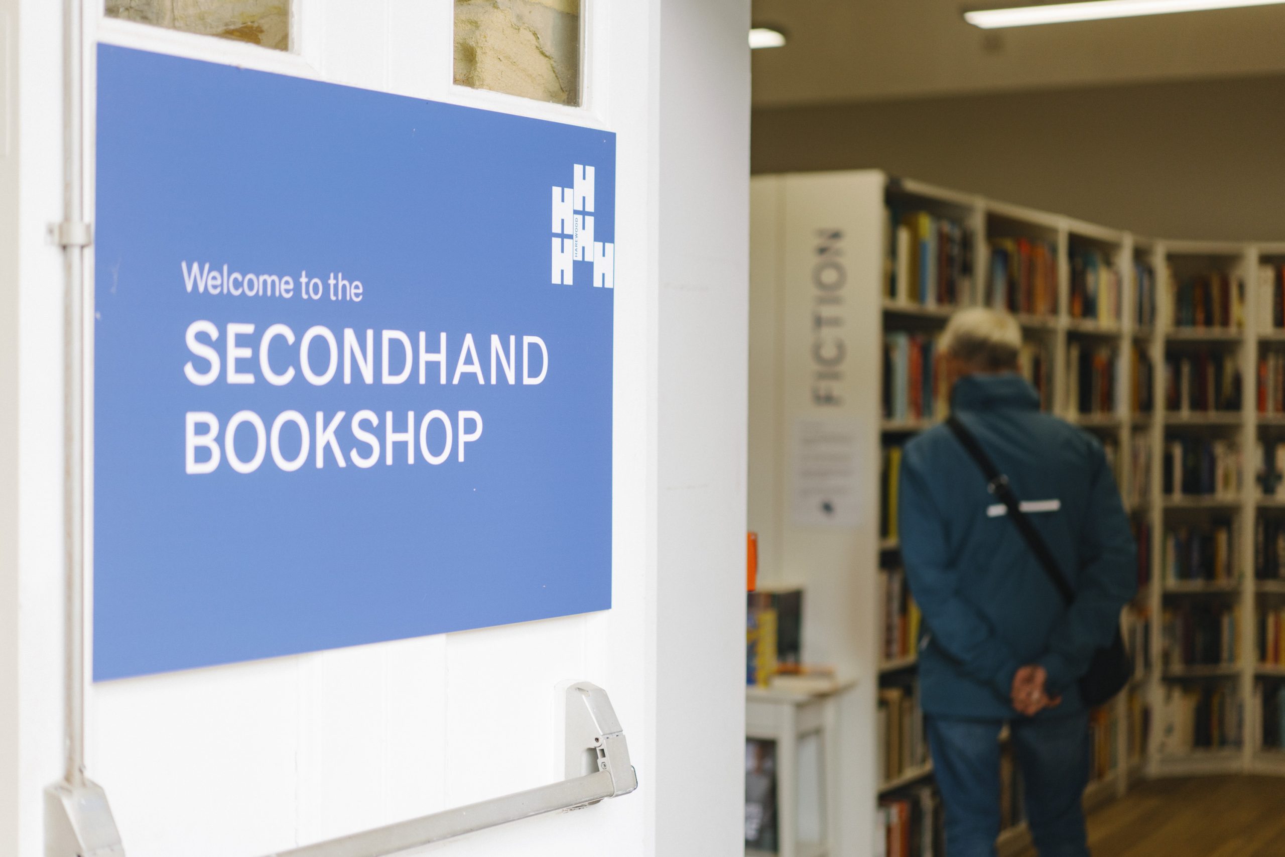 A vibrant blue sign which reads 'Welcome to our Secondhand Bookshop', hangs on an open door as a man browsers the shelves of books.