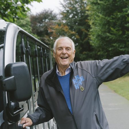 A very happy, playful, caring and helpful person wearing a volunteer Harewood fleece decorated with a long service award pin badge. The person is stood with arms wide in a happy manner, in front of a small white shuttle bus.