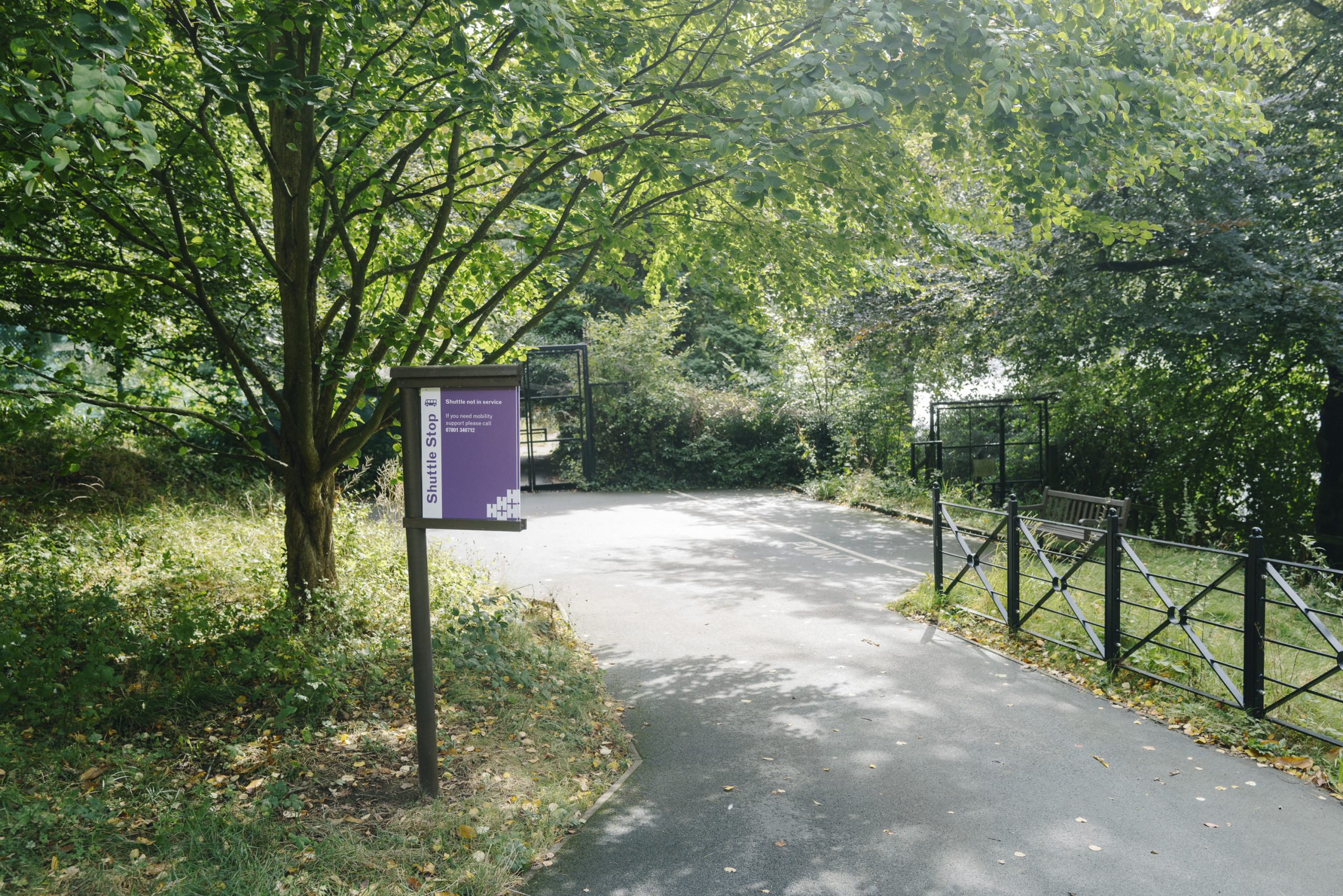 Pathway surrounded by lush greenery, with a purple "Shuttle Stop" sign on the left. A bench and fence line the right side, creating a peaceful setting.