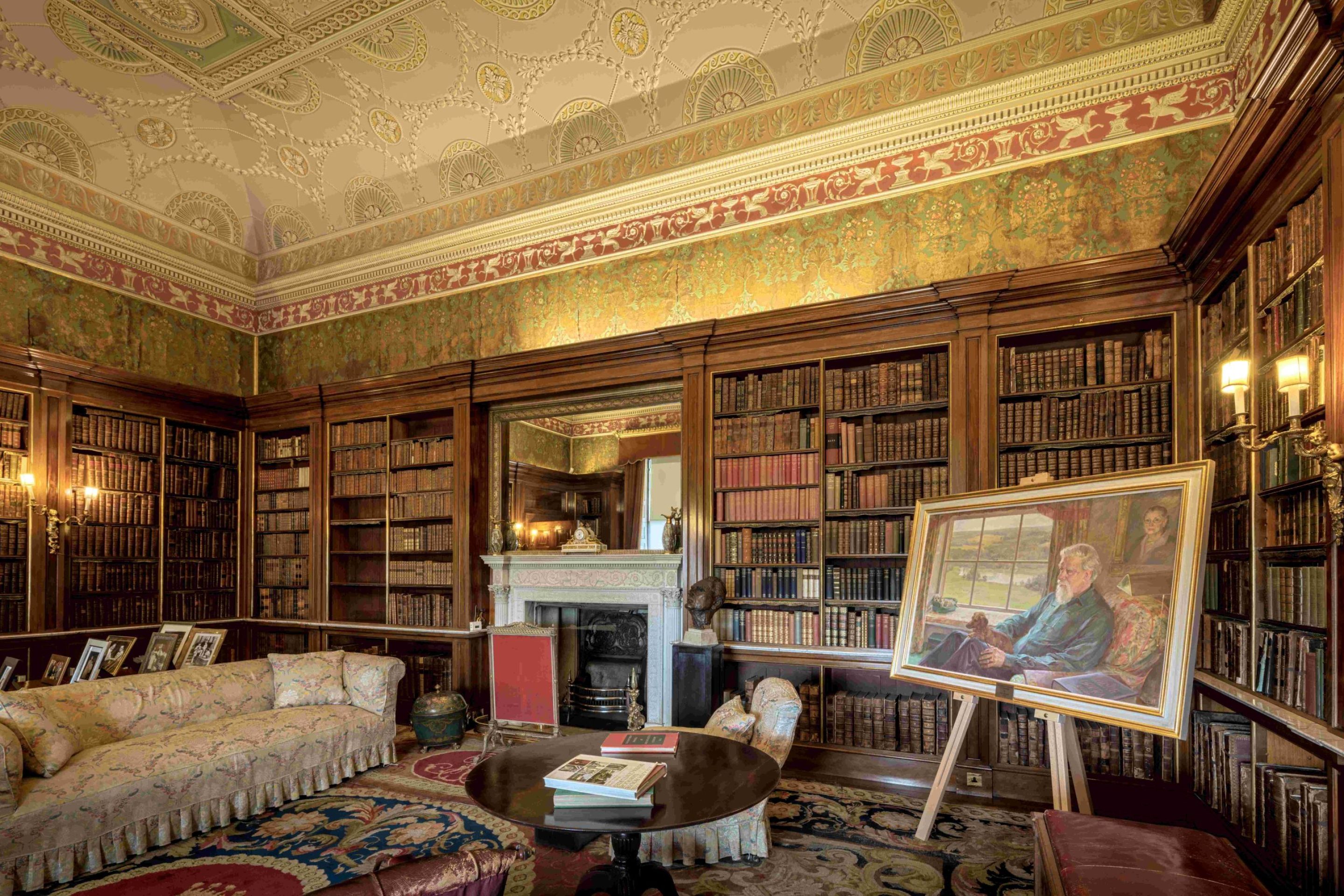 A large room with a decorative plasterwork ceiling painted in pastel colours. The walls are entirely encased in large mahogany bookshelves. There are various pieces of furniture and a portrait on an easel in one corner.