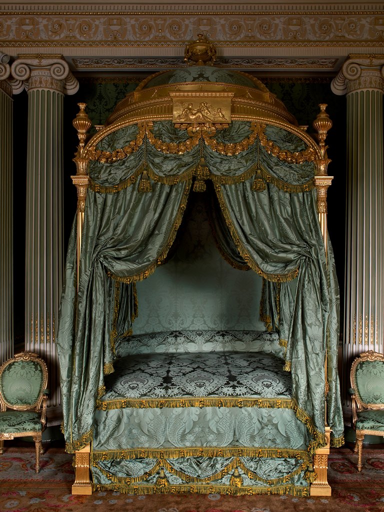 A large four-poster bed with a domed canopy. The frame is entirely gilded and upholstered in rich green damask.