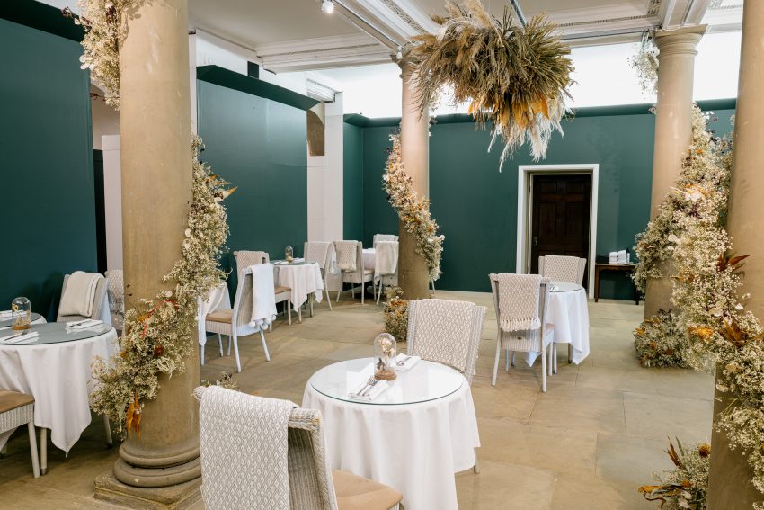 Terrace Cafe indoor seating area with round tables generously spaced apart. Blankets drape over the back of each seat with the tables dressed with dried flowers. Ornate pillars decorated with swirling dried flower arrangements.