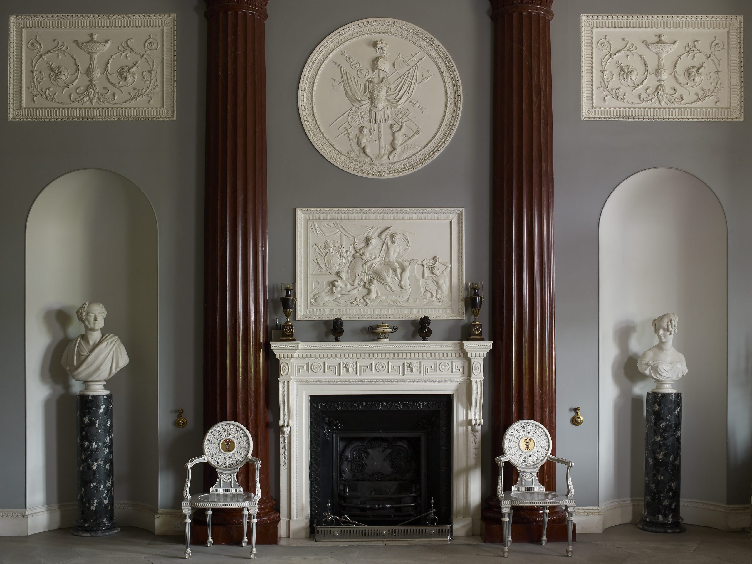 An elegant neo-classical fireplace adorned with a marble mantel, paired with two matching chairs designed by Thomas Chippendale. Either side of the fireplace, two marble busts stand on marble plinths. The Entrance Hall is designed to look like the inside of a classical temple.