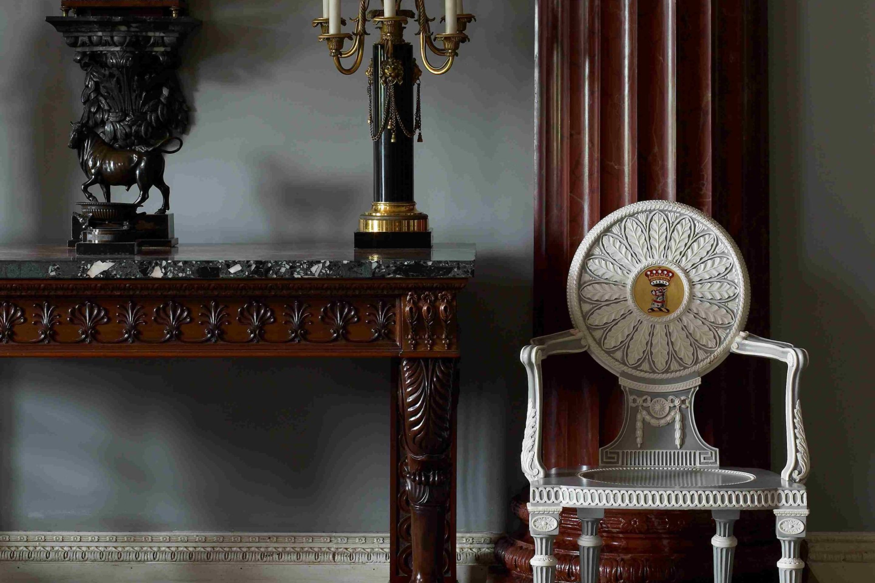 An elaborate white and blue painted chair next to a carved mahogany side table upon which stands a bronze lamp.