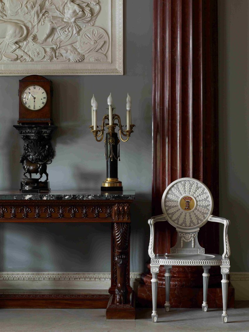 An elaborate white and blue painted chair next to a carved mahogany side table upon which stands a bronze lamp.