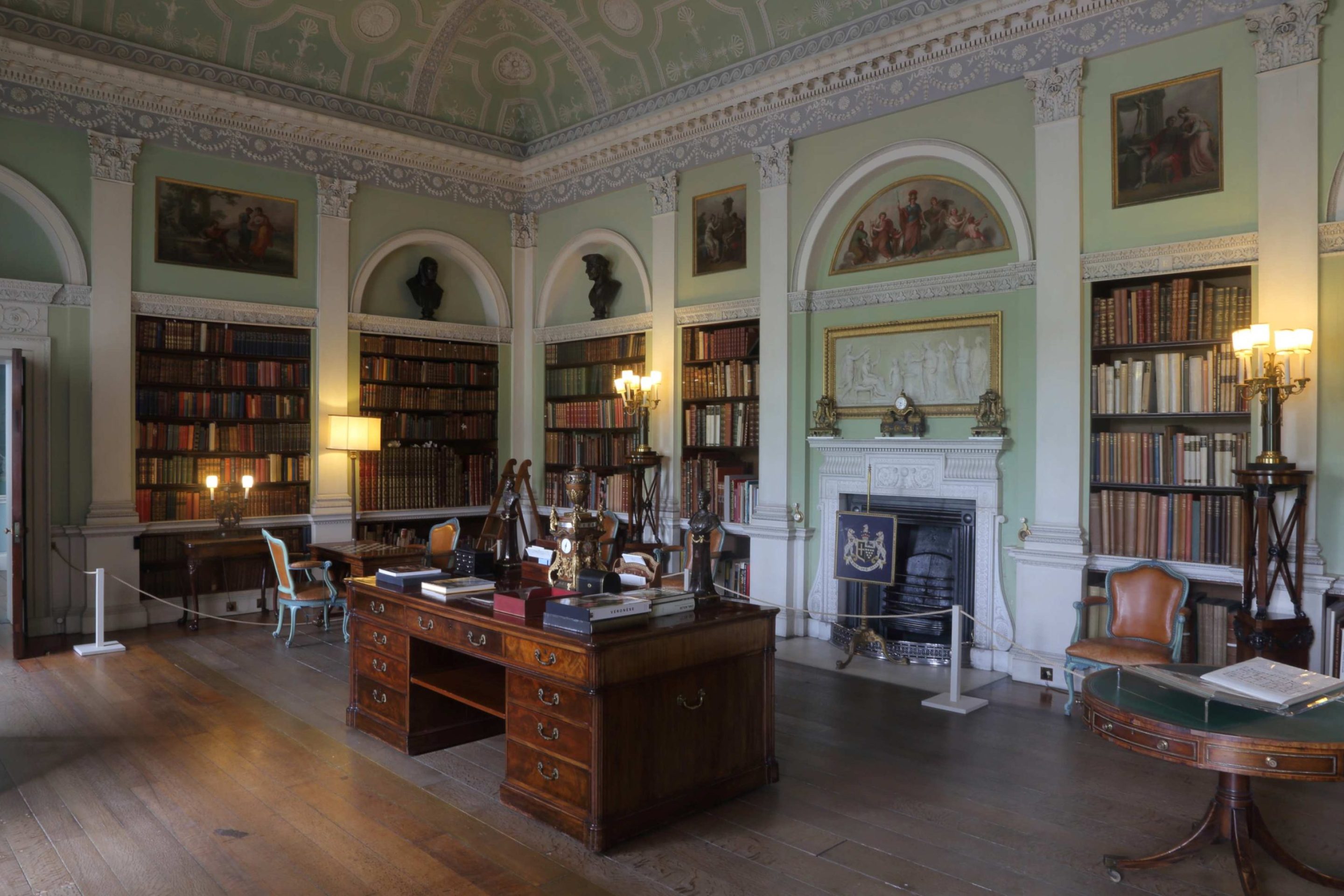 A library with bookshelves divided by classical columns. A large mahogany desk stands in the centre of the room.