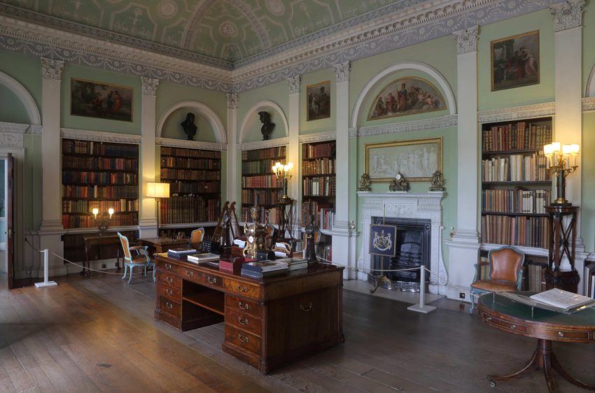 A library with bookshelves divided by classical columns. A large mahogany desk stands in the centre of the room.