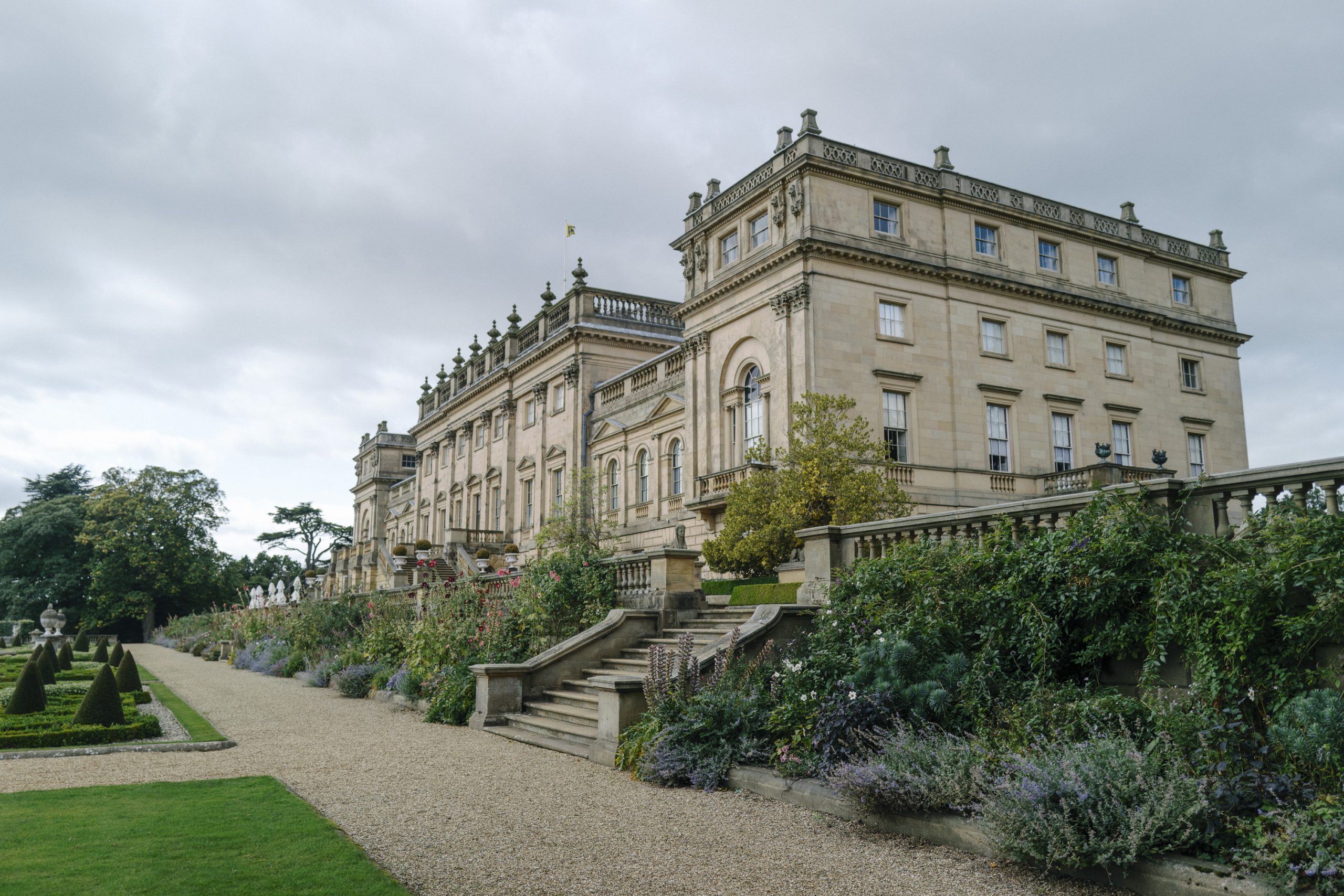 Grand Georgian country House with elegant architecture, surrounded by lush gardens and a manicured pathway, under a cloudy sky.