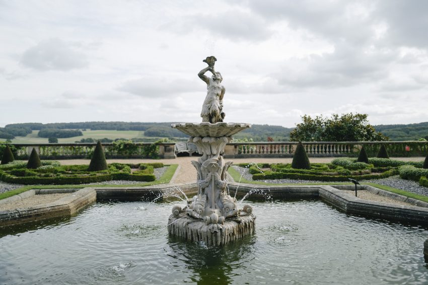 A stone fountain of a man blowing water through a horn into the sky with fish heads spraying water into the pool below creating ripples under a clouded sky.