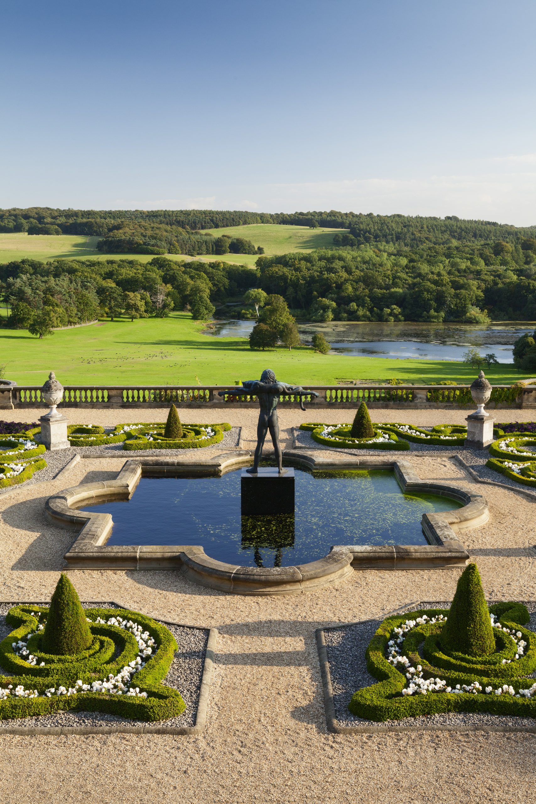 Clear blue skies sit above the rolling Capability Brown landscape with trees dotted across the horizon. Orpheus stands proudly in the middle of an ornate pool of water on the Victorian Terrace.