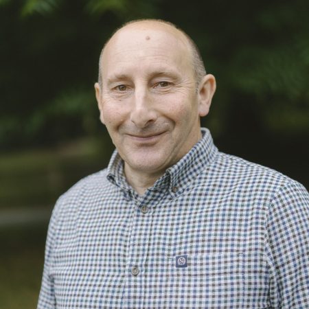 Man wearing a blue and brown checkered long sleeve shirt, smiling warmly against a lush green background.