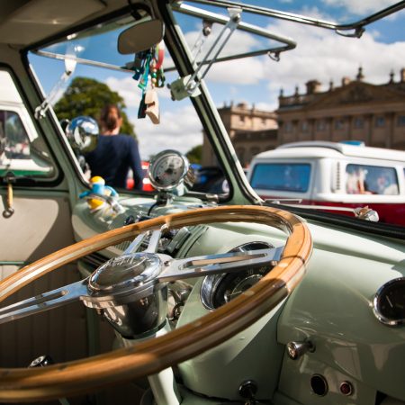 Sage green vintage Volkswagen Beetle parked in front of Harewood House. The dashboard is so clean it's sparkling in the sunshine whilst the wooden steering wheel reflects the golden glow. People wander around in the background, looking at the other vintage Volkswagen cars.