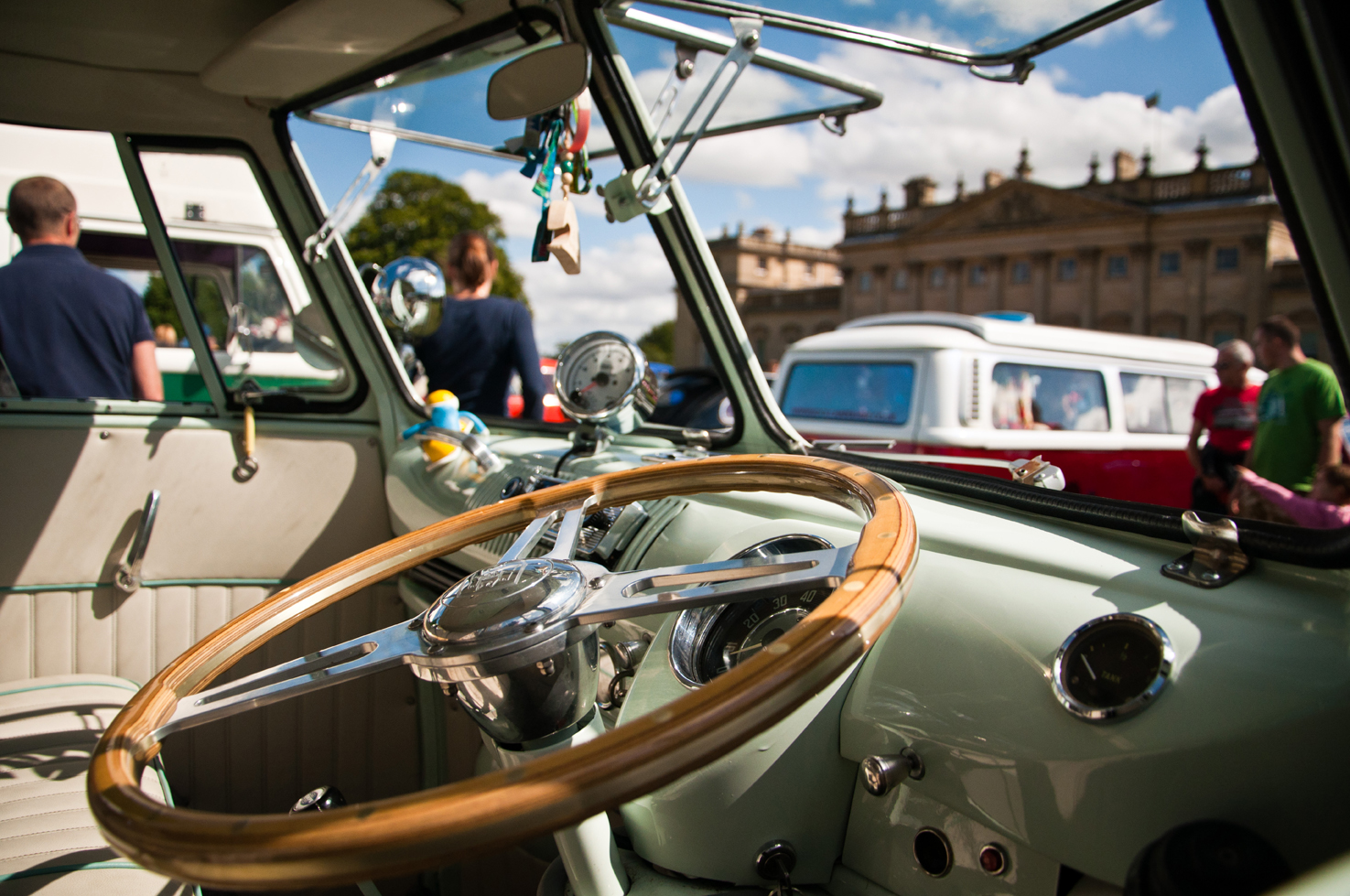 Sage green vintage Volkswagen Beetle parked in front of Harewood House. The dashboard is so clean it's sparkling in the sunshine whilst the wooden steering wheel reflects the golden glow. People wander around in the background, looking at the other vintage Volkswagen cars.