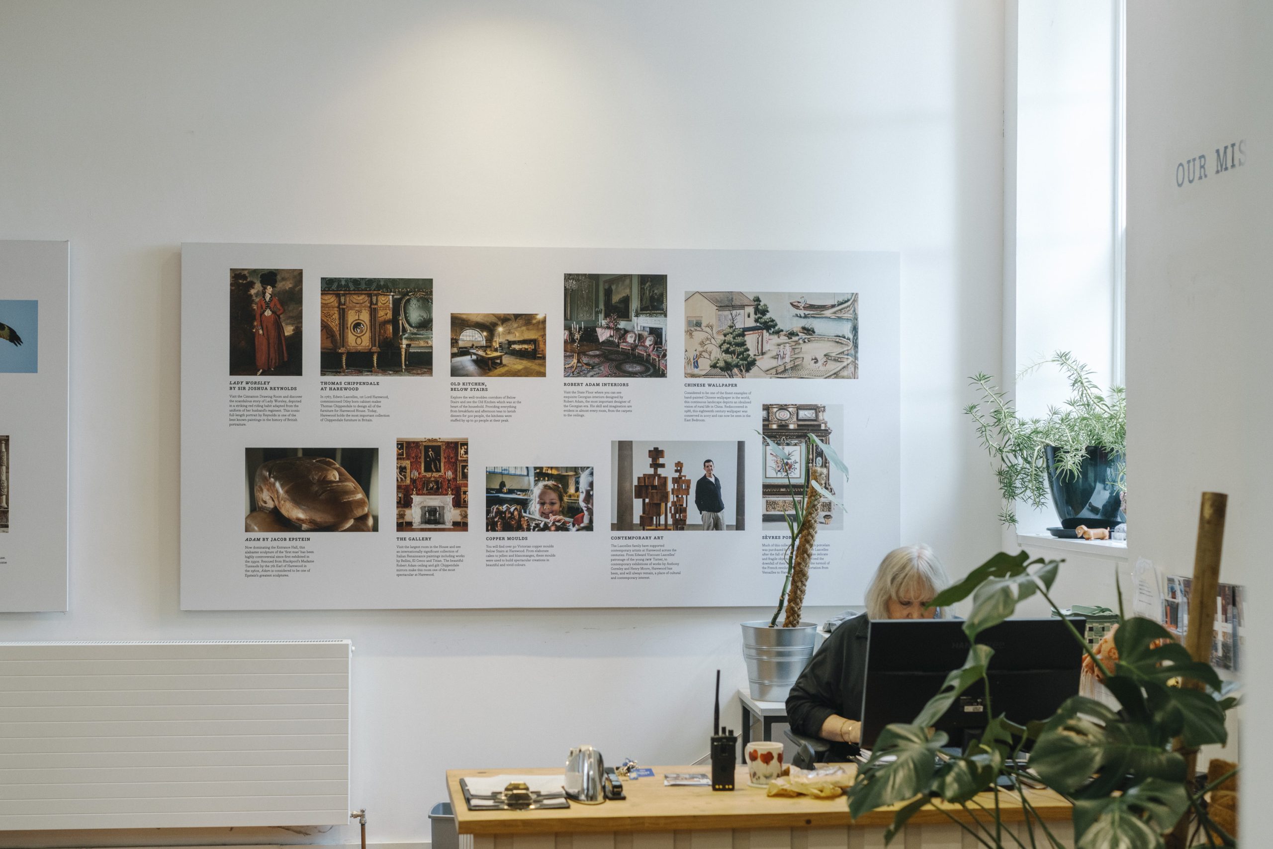 A women with short blonde hair sits behind a desk surrounded by green plants. A radio stands on the desk next to a steaming cup of coffee. Behind the desk, upon the wall is photos of Harewood's collections including Chippendale furniture, British Portraiture, Chinese handpainted wall paper and scuptures.