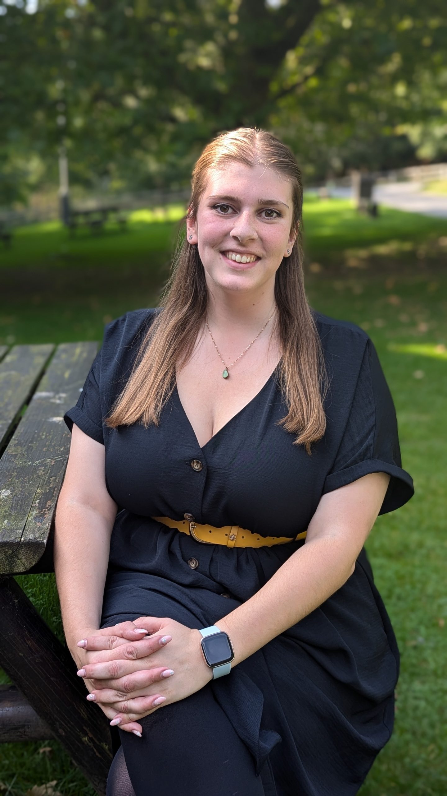 A smiling person with long, straight brown hair, wearing a black top with a green teardrop necklace sat on a picnic bench in a park.