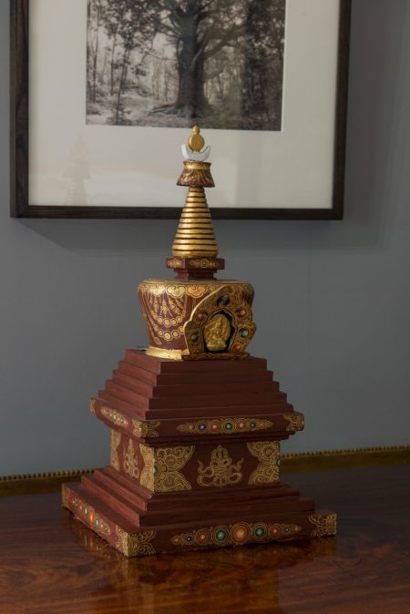 A model of a Buddist stupa with a square base and tiered steps leading towards a conical top. It is painted a dark brown with gilded patterns.