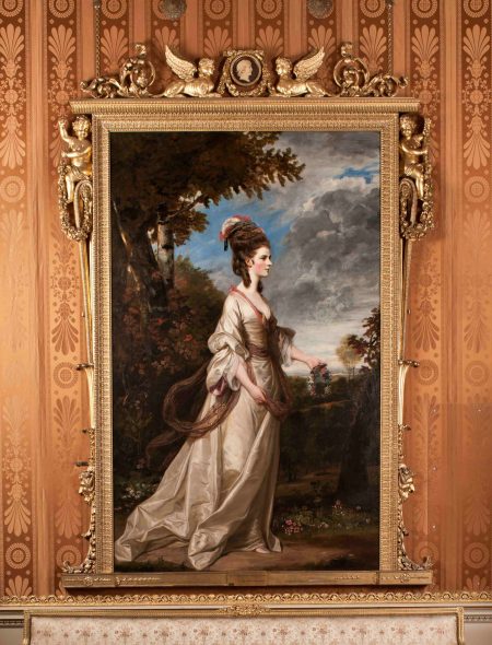 A full length oil painting of a white woman in her early 20s. The sitter, Lady Harrington, stands before a wooded grove and a bright blue sky. She wears a flowing white gown and beehive-style hair. She holds a wreath of flowers.