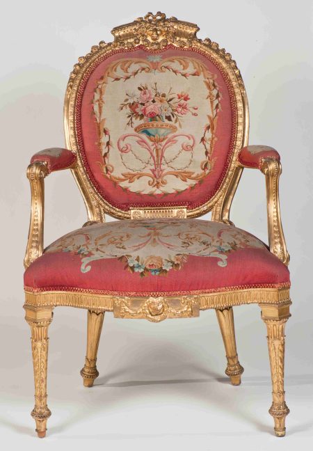 A eighteenth-century armchair with elaborately carved decoration and gilded surface. The seat, back and arms are upholstered in a pink floral tapestry.