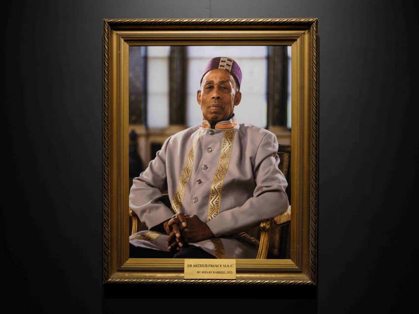 A half-length photographic portrait in a gilded frame of a Black man in his 80s. The sitter, Arthur France, is shown seated on a gilded chair. He wears a lilac jacket with gold embroidery and a purple pill-box-style hat.