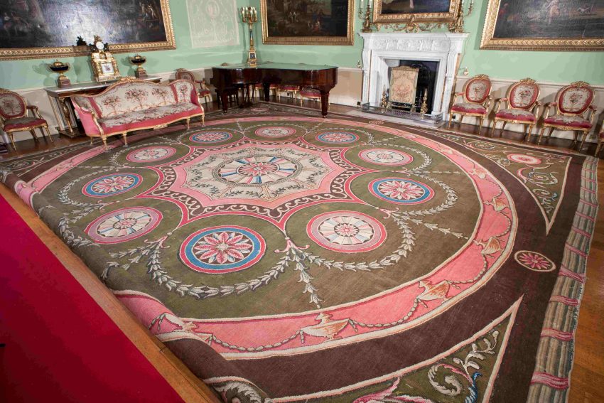 A large square carpet, rolled on one side, laid on the floor of a historic interior. Its colour scheme is mainly pink and green and highly patterned.