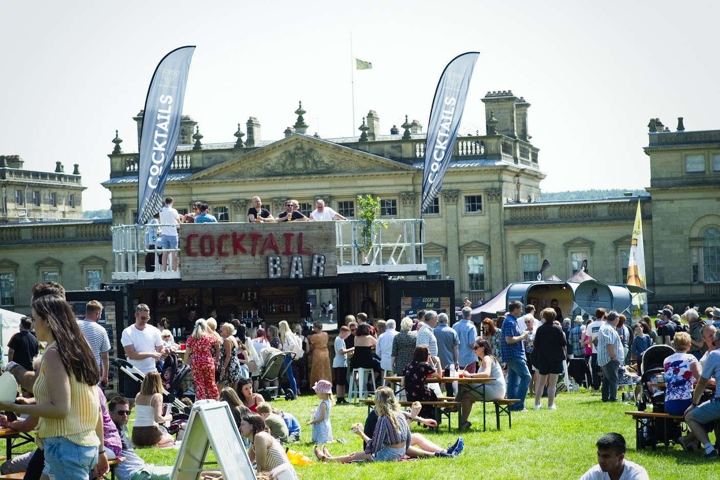 A group of people relax on the grass on a variety of seating in front of a large building, enjoying a sunny day outdoors and eating various foods. A cocktail bar sits in front of the building with people enjoying their drinks