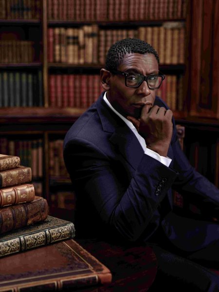 A half-length photographic portrait of a Black man in his 50s. The sitter, David Harewood, is shown seated with his elbow on a table with his hand raised to his mouth. He wears a bark blue suit and white shirt. He sits in front of mahogany bookshelves, and a pile of books is placed to his left.