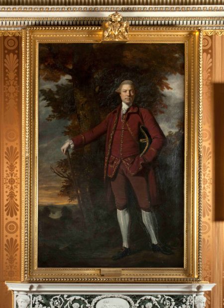 A full-length oil portrait of a white man in his 50s. The sitter, Edwin Lascelles, is shown standing in the Harewood estate before a great oak tree and Harewood House in the distance. He stands with one arm on a cane and the other in his pocket, wearing a deep plum suit typical of the 18th century with gold embroidery.