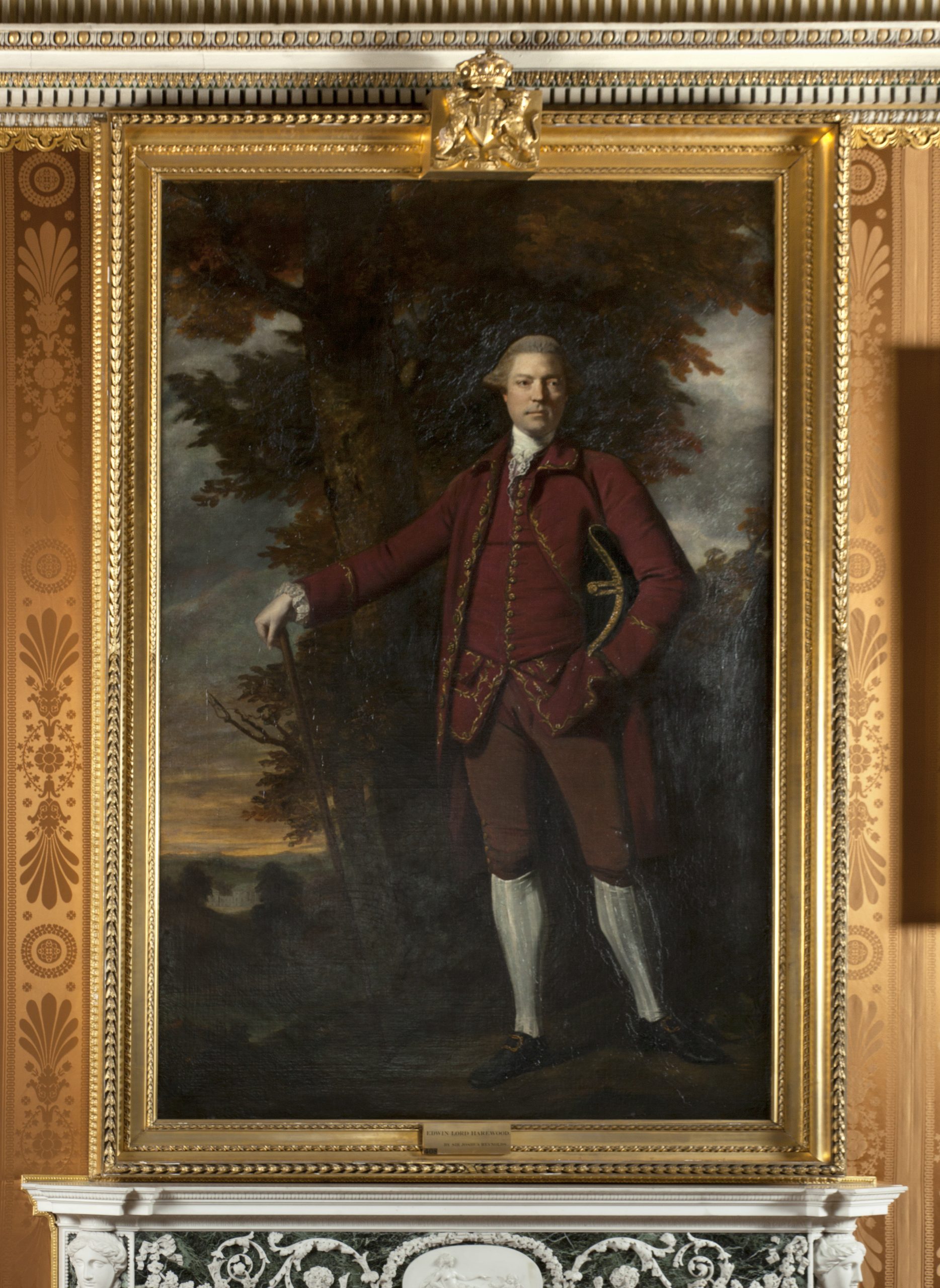 A portrait of Edwin Lascelles, a tall man with a foreboding stare. He wears a smart outfit, typical of 18th century. A red full-skirted knee-length coat, matching knee breeches and long waistcoat, with a white linen shirt and white linen underdrawers. He stands proudly holding a cane out to the side. The portrait is inside a gilt frame above a marble fireplace.