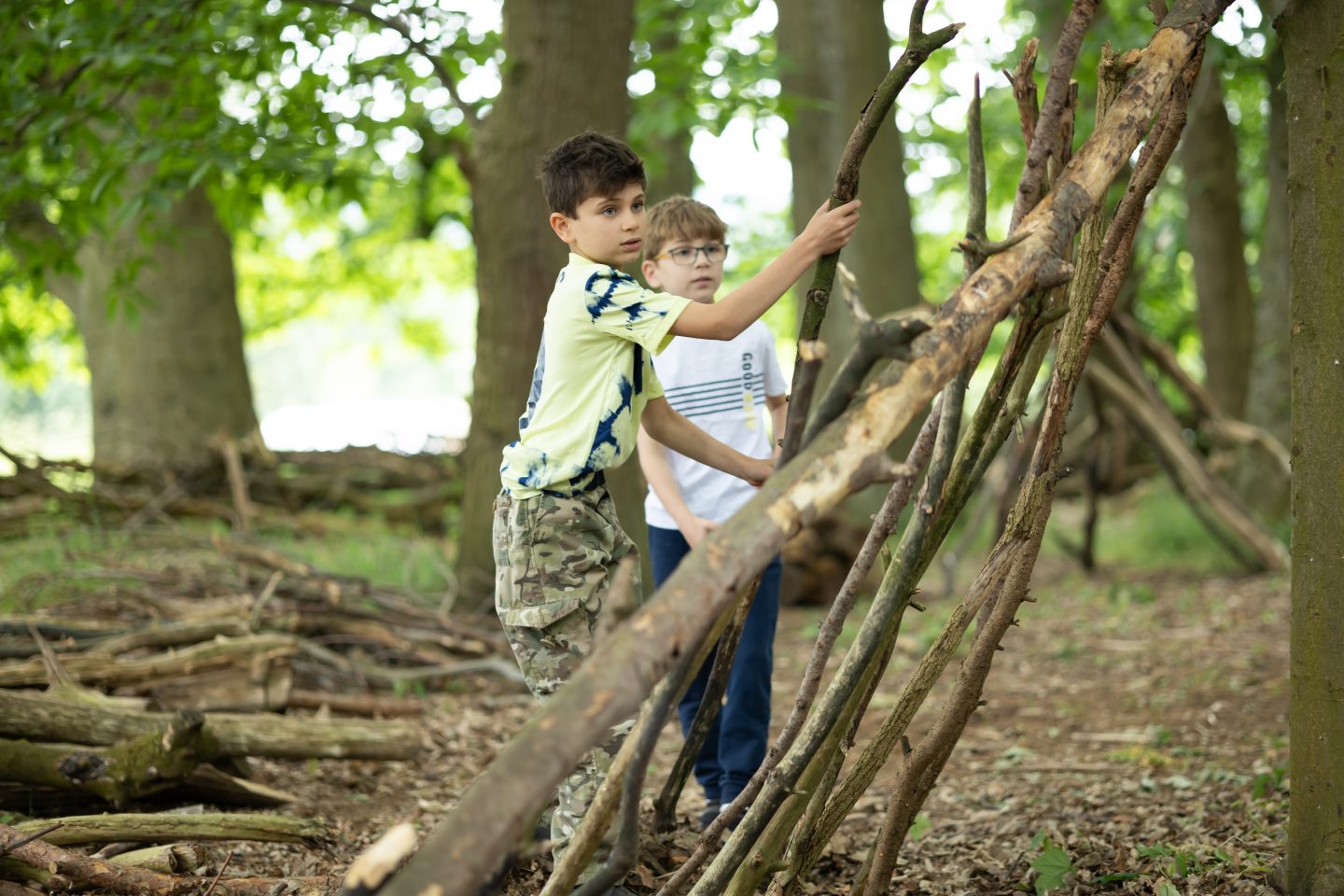 Two boys outdoors building a stick shelter in a Harewood woodland, amid tall trees and leafy greenery. The scene conveys teamwork and playful curiosity.