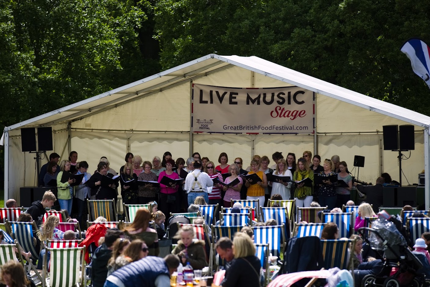 A large group of people, seated in striped deck chairs in front of a tent. Inside the open fronted tent is a group of people singing in front of a 'live music' banner