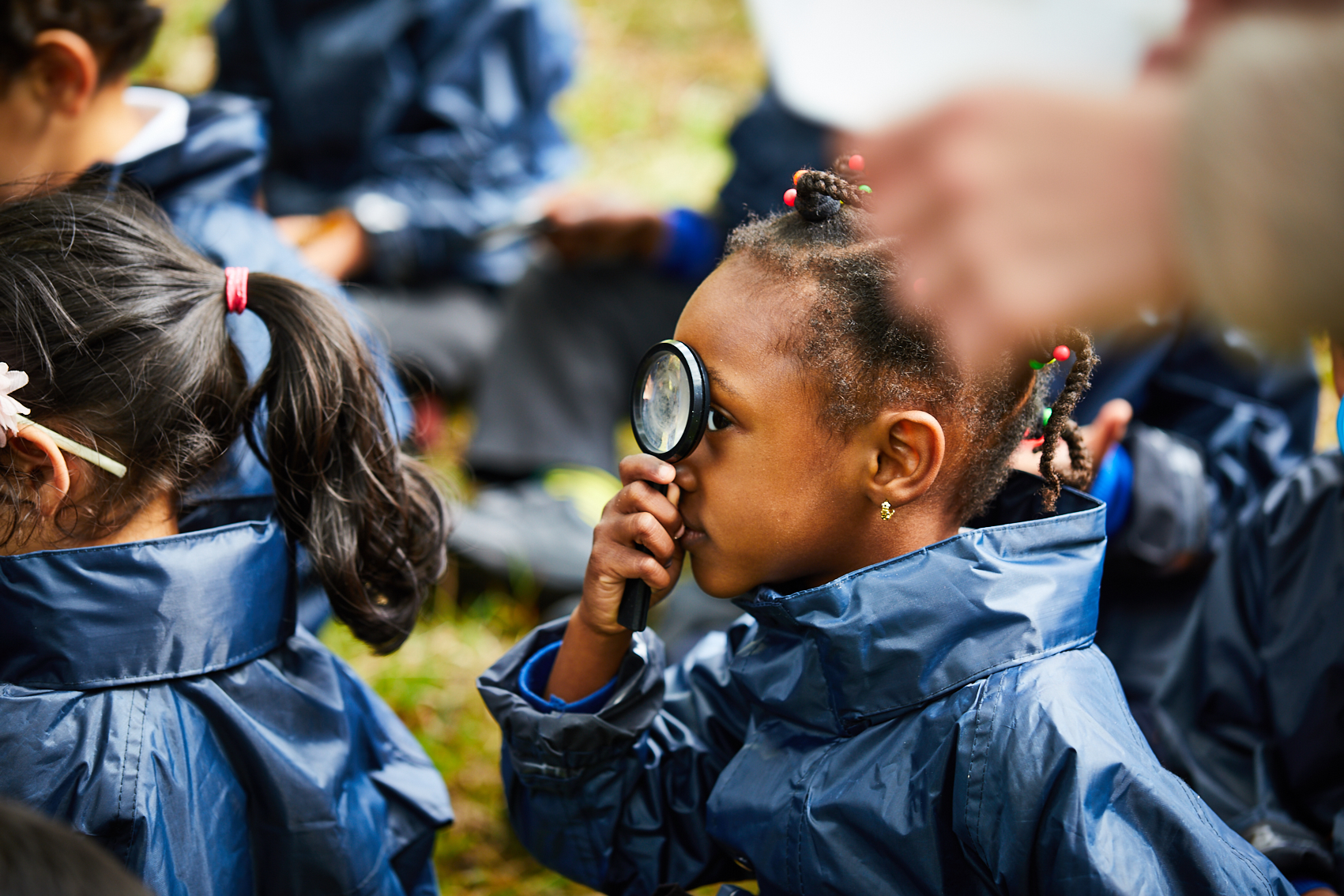 A young girl in a blue raincoat peers through a magnifying glass, focusing intently. She is surrounded by other children in similar attire outdoors.