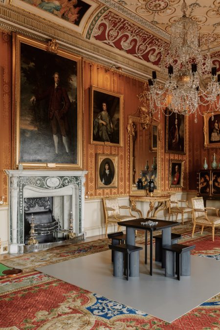 A large historic interior with cinnamon-coloured damask walls and various portraits. In the centre of the room is a black, square-shaped games table with aluminium dominoes.
